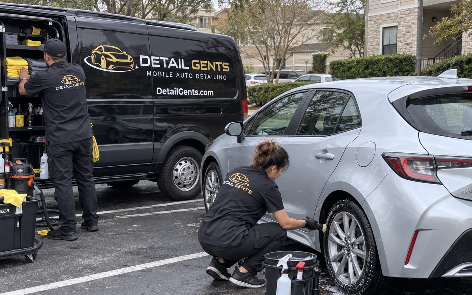 A Detail Gents detailer washing the wheels of a silver hatchback in an apartment community parking lot