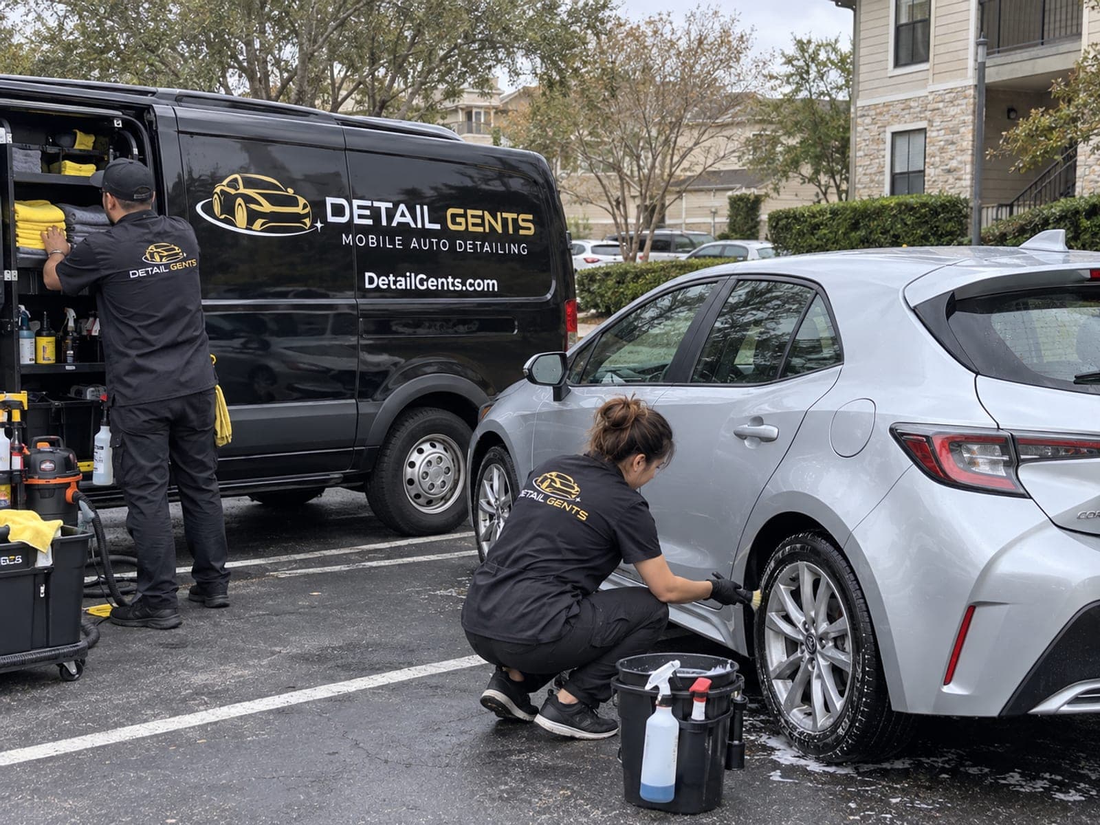 A branded Detail Gents van parked in a bay at an apartment community while a detailer works on a car alongside